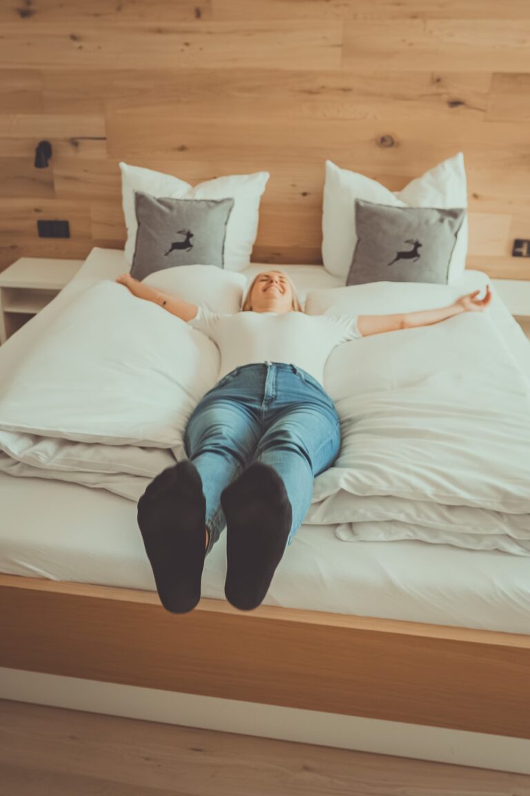 Eine glücklich lächelnde Frau liegt entspannt mit ausgestreckten Armen auf einem großen Doppelbett im Hotel Mariandl am Berg. Der Raum besticht durch eine moderne Holzwand, helle Bettwäsche und stilvolle Kissen mit Hirschmotiv.